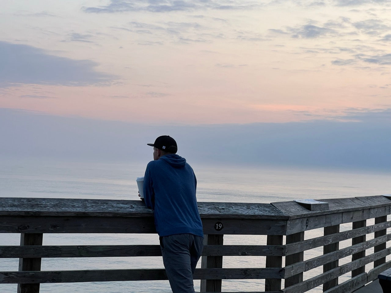 Man standing at pier railing looking into a calm sunrise over the ocean