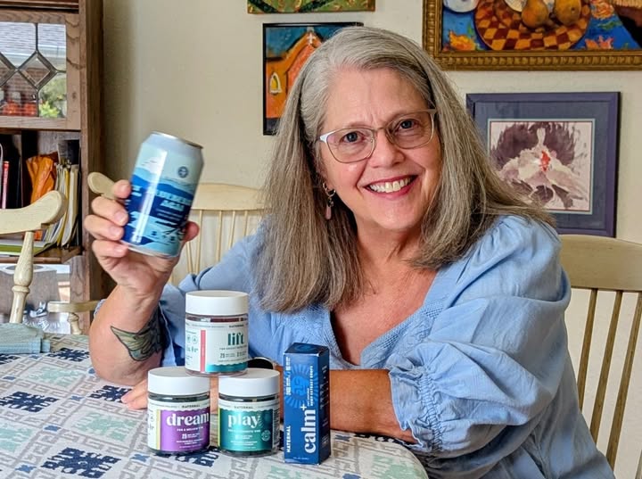 A Naternal customer at her kitchen table, smiling, holding a Blueberry Acai seltzer with Lift, Dream, Play and Calm+ in front of her